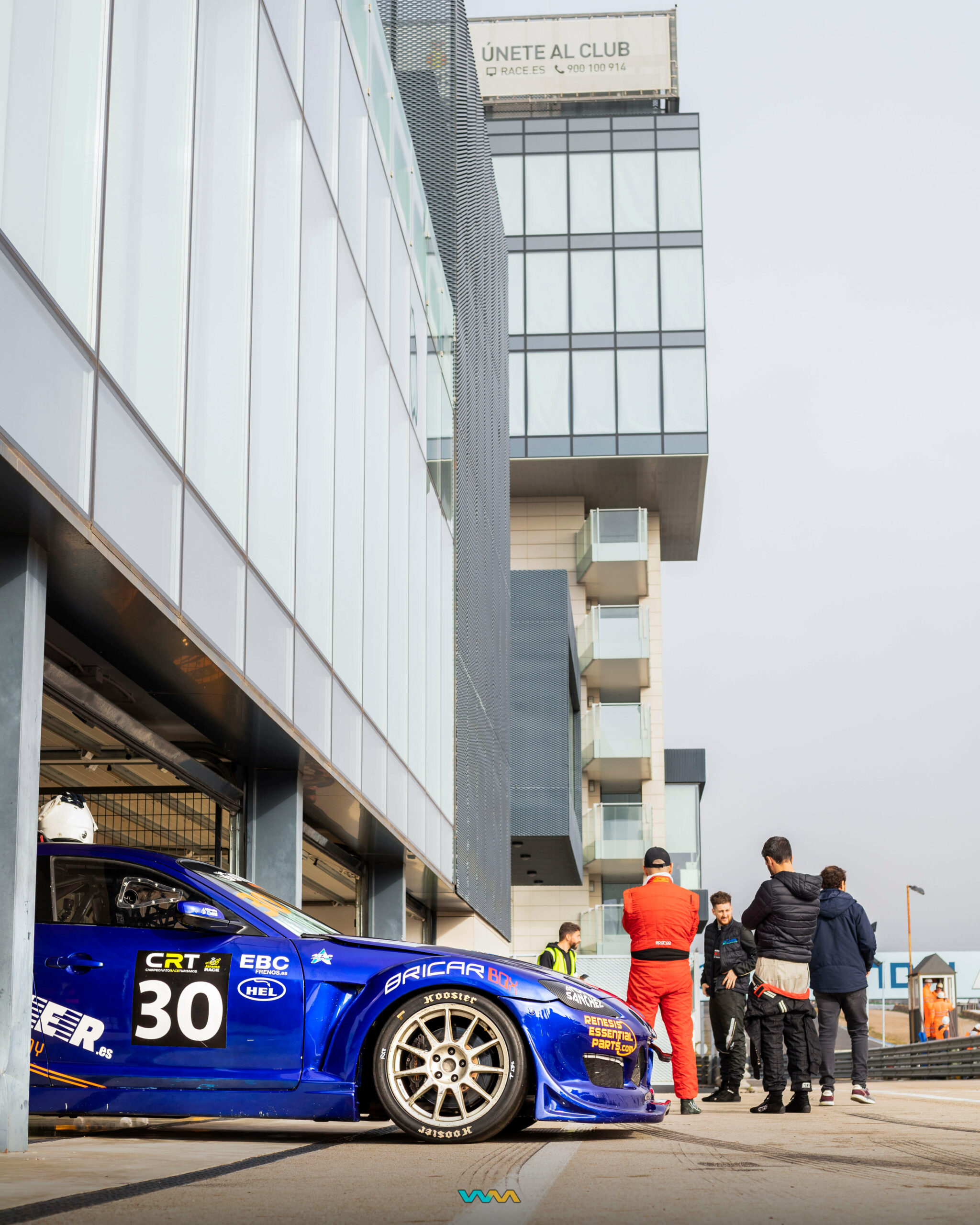 Mazda RX8 en el box con mecánicos y pilotos cerca de él y la torre del Jarama de fondo en el Campeonato Race de Turismos 2023. Fotografía de Warmedia