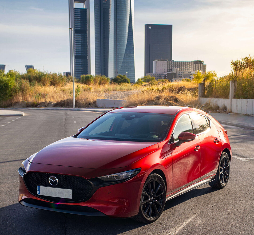 Mazda 3 rojo fotografiado al atardecer con las Cuatro Torres Business Area de Madrid al fondo, en una imagen con estilo Warmedia: luz cálida, encuadre limpio y enfoque comercial para venta de vehículos