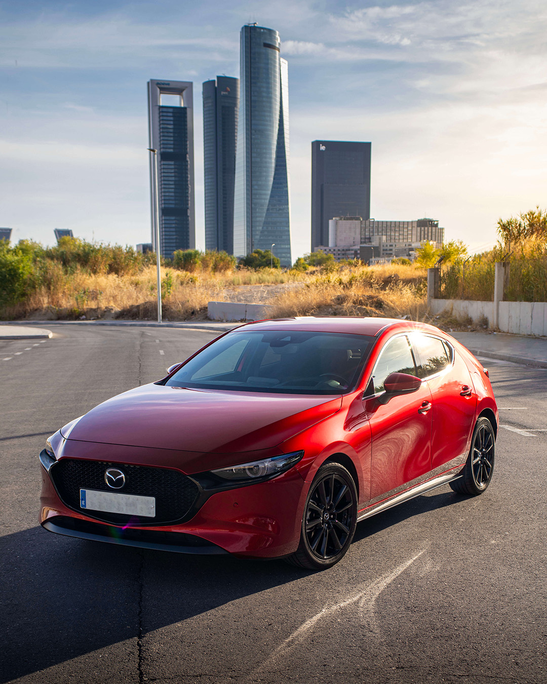 Mazda 3 rojo fotografiado al atardecer con las Cuatro Torres Business Area de Madrid al fondo, en una imagen con estilo Warmedia: luz cálida, encuadre limpio y enfoque comercial para venta de vehículos